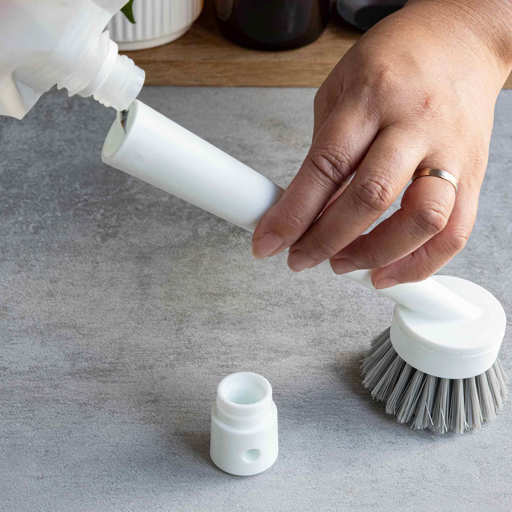 Person using a white bottle to fill a gray scrubber with liquid on a gray surface. Simply Clean Scrub Brush - White