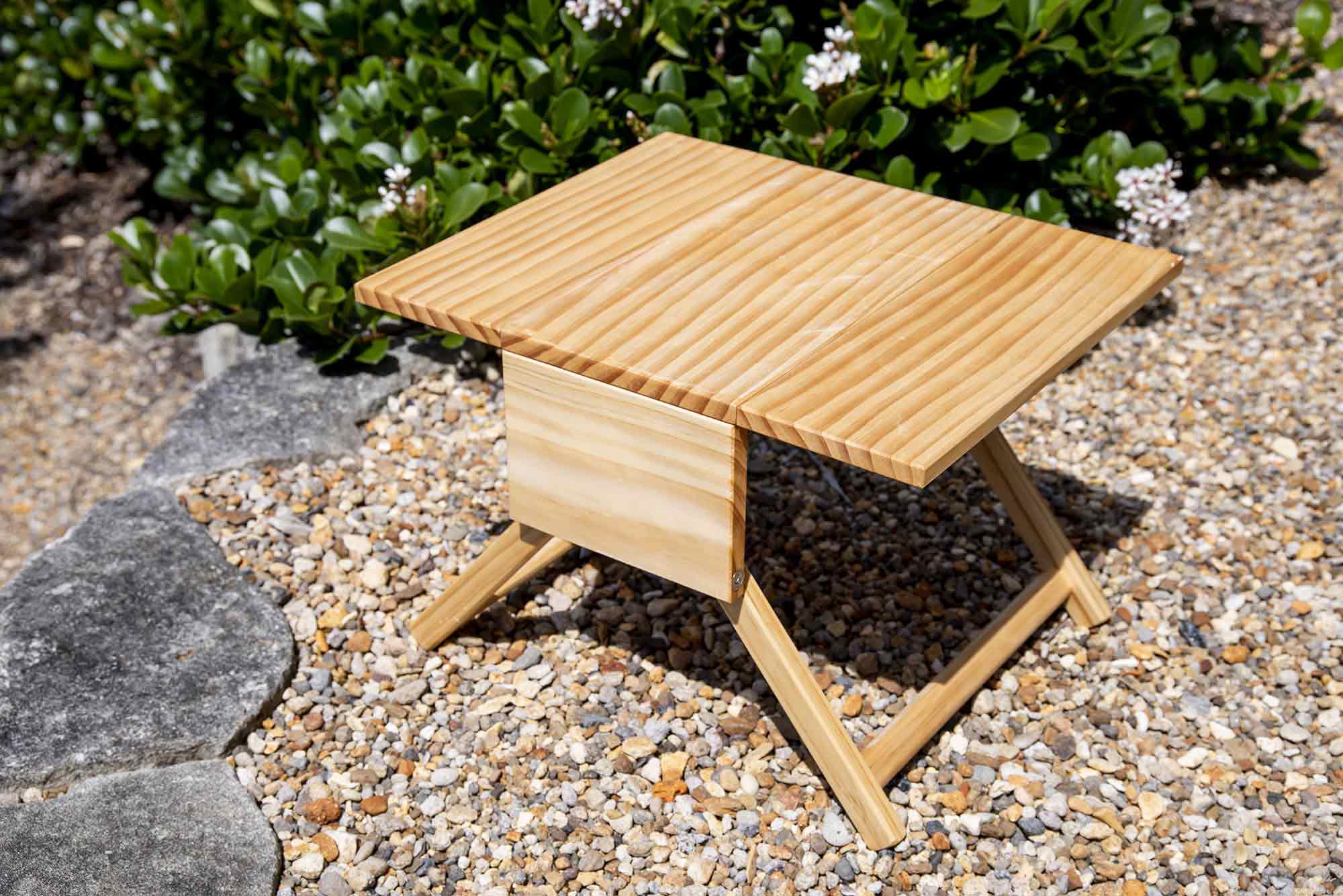Wooden folding table on a gravel surface with greenery in the background. Victoria James Home Storage Box VJA Convertible