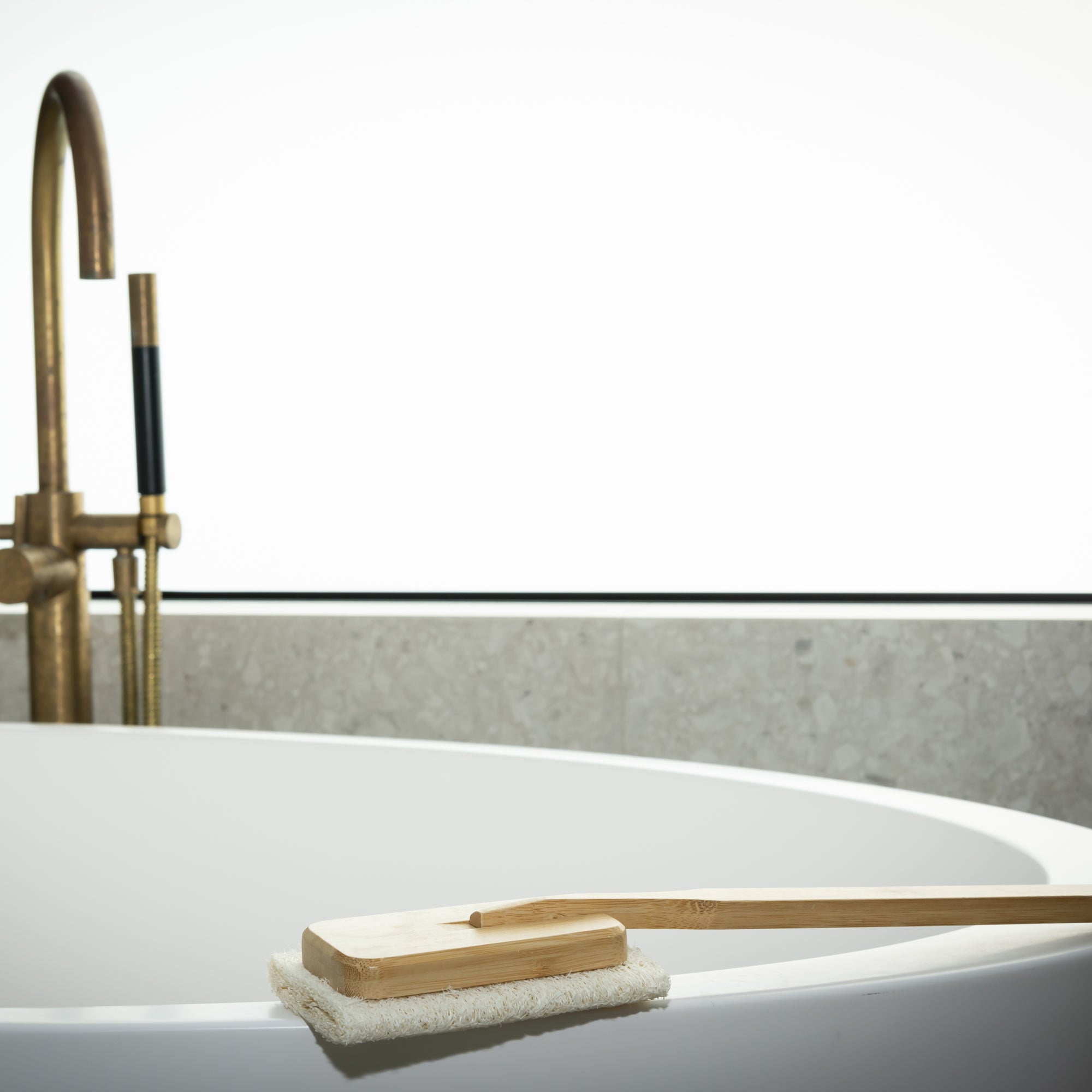 Bath brush with a wooden handle and white bristles on a bathtub, with a blurred background of a bathtub and faucet. Victoria James Body Loofah Brush - Square
