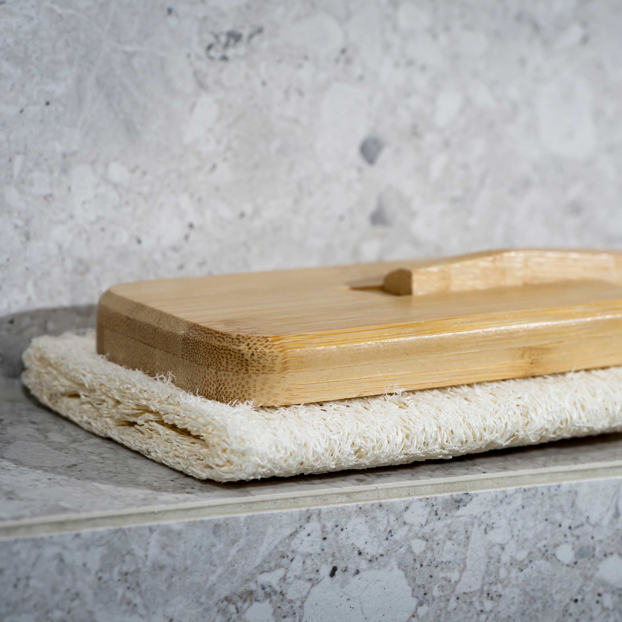 Bamboo cutting board on a stone surface with a textured gray background. Victoria James Body Loofah Brush - Square