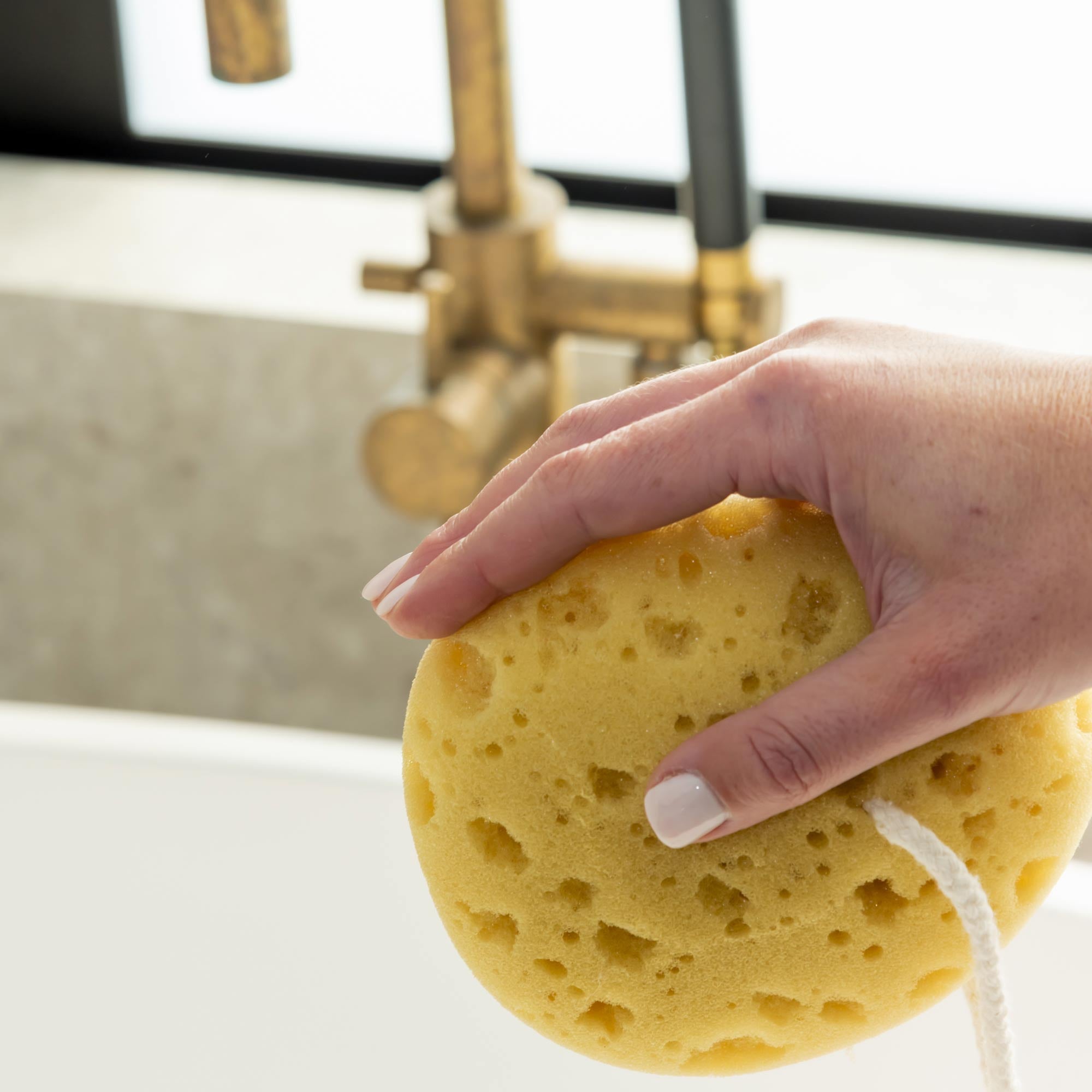 Hand holding a yellow sponge over a sink with a gold faucet. Victoria James Bath Sponge