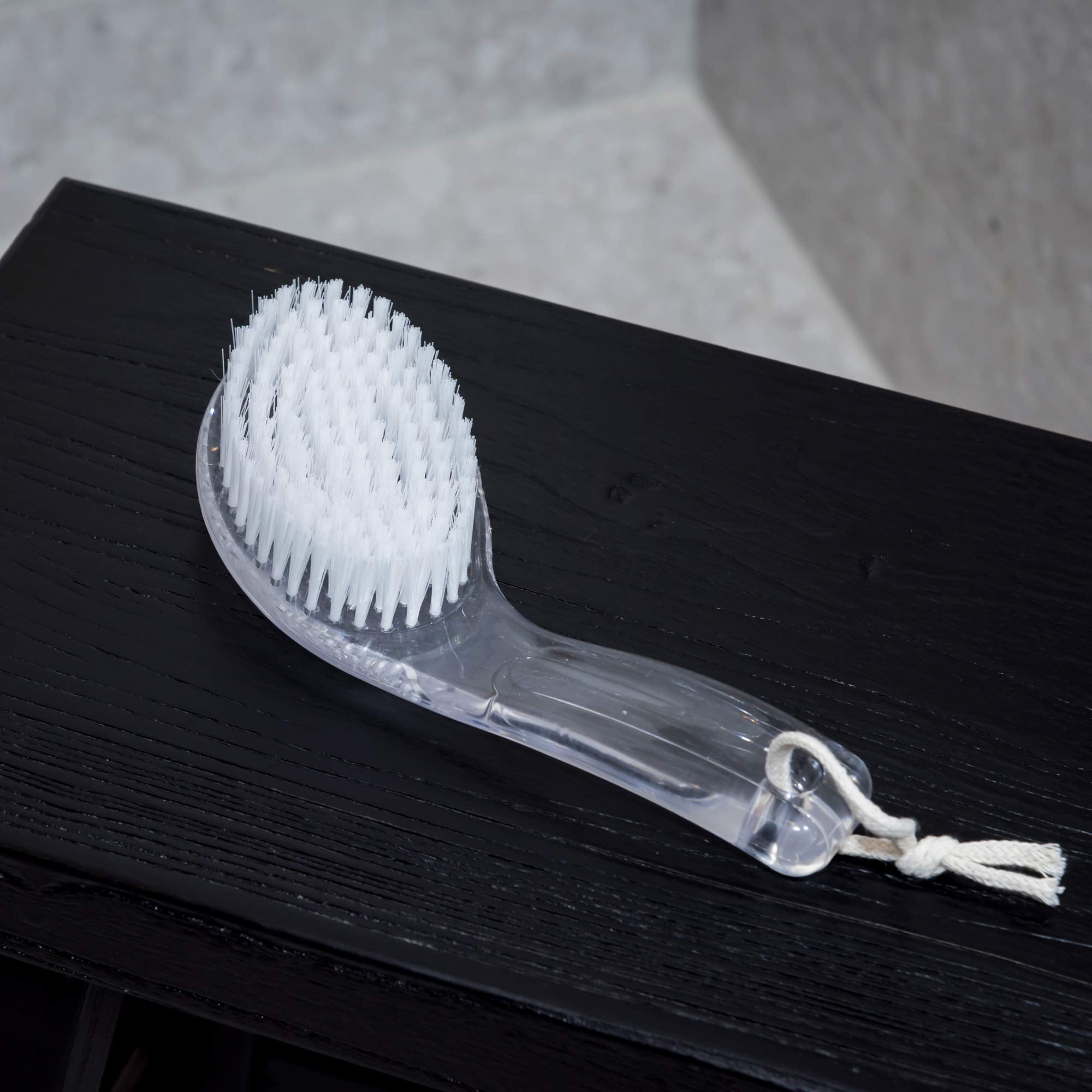 White scrubbing brush with a clear handle on a dark surface. Victoria James Body Bath & Shower Brush