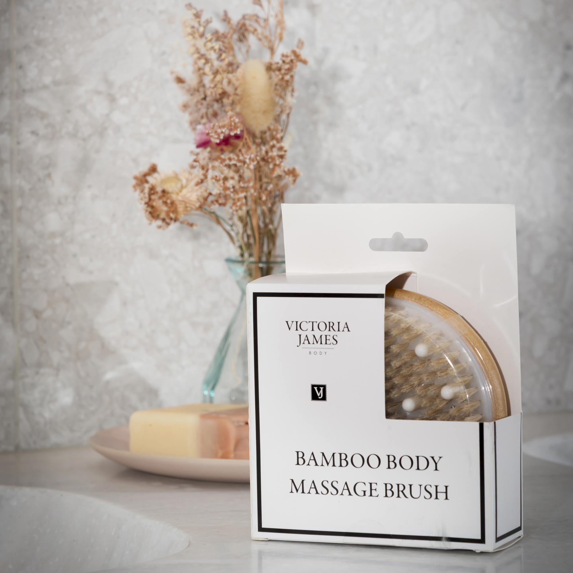Victoria James Bamboo Body Massage Brush packaging with a blurred background of candles and flowers. Victoria James Massaging Body Brush
