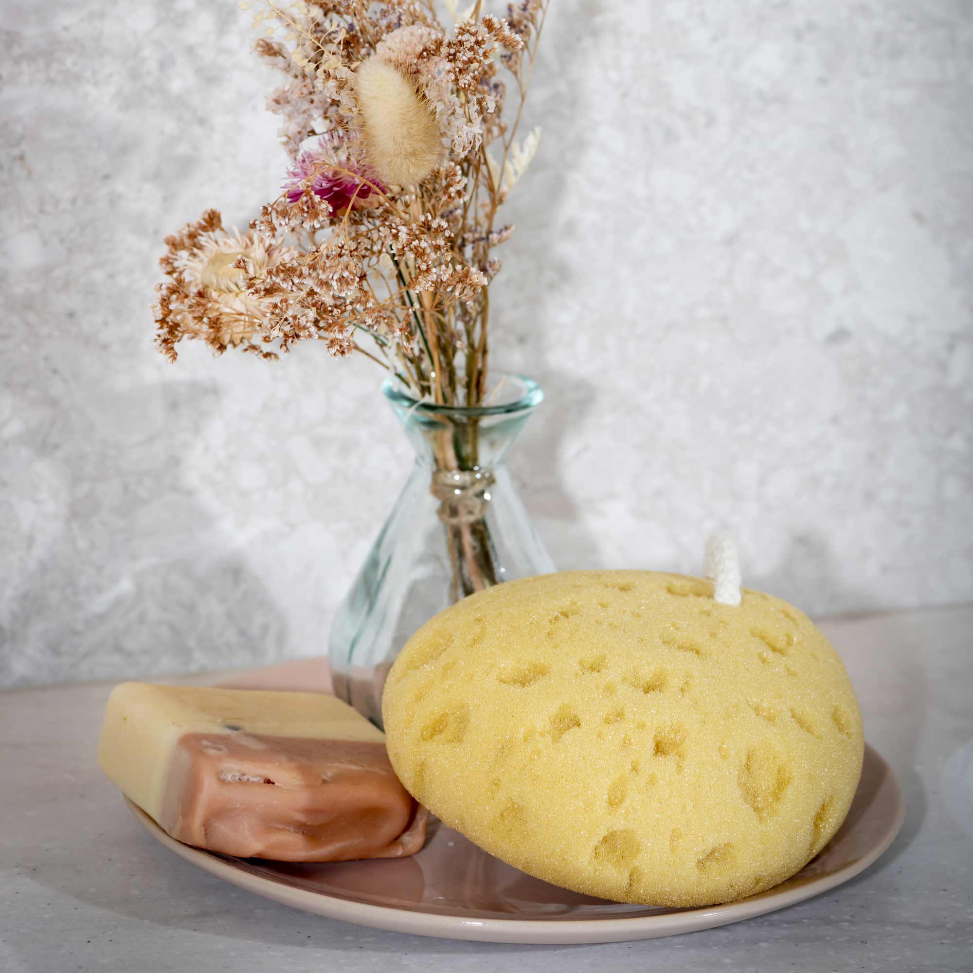 Yellow sponge candle on a plate with a bar of soap and a vase of dried flowers in the background. Victoria James Bath Sponge