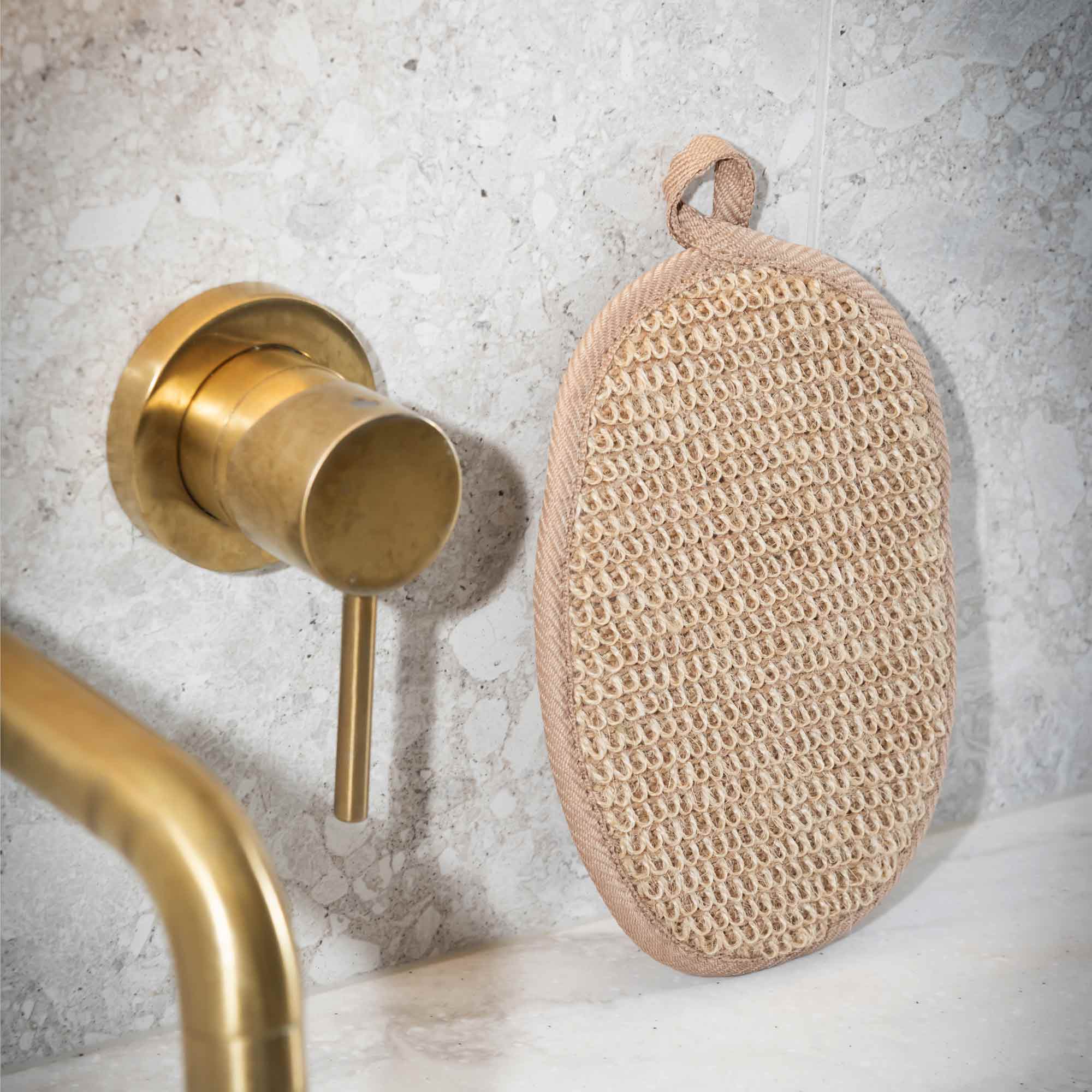 Brass faucet and loofah on a textured wall. Victoria James Body Exfoliating Pad